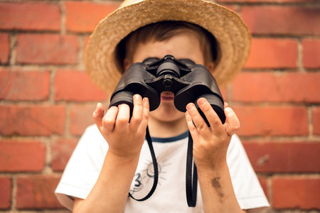 Boy in a straw hat looks in binoculars on the background of a brick wall.