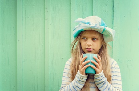 Cute little girl stands near a wall in a turquoise hat and holding a cup. Space for textの写真素材