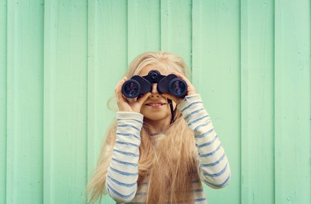 Cute little girl stands near a turquoise wall and looks binoculars. Space for text.Negative speace.の写真素材