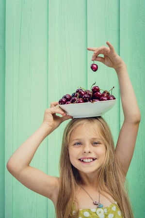 Beautiful blond girl on a background of turquoise wall holding a plate with cherries on her head. Space for textの写真素材