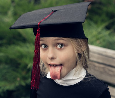 Beautiful blond girl in a school uniform and graduation cap on his head sticks out his tongue as Albert Einstein. Education.の写真素材