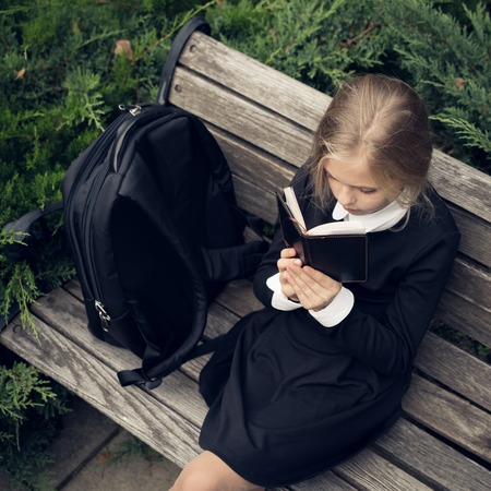 Beautiful blond girl in a school uniform sits on a park bench and reading a book. School uniform.Educationの写真素材