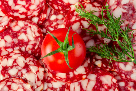 A large ripe red tomato and a branch of dill lies on a blurred background of sliced salami. Close-up.の写真素材