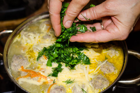 Preparation of homemade soup with meatballs and vegetables in a metal saucepan. Hands pour greens...