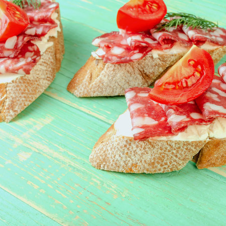 Fresh tasty salami sandwiches. Appetizing sandwiches with smoked sausage, cream cheese and tomatoes. On blue wooden background. Square. Close-up.の写真素材