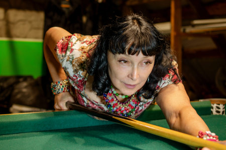 Beautiful adult woman concentrates aiming billiard cue on the green billiard table. The concept of...