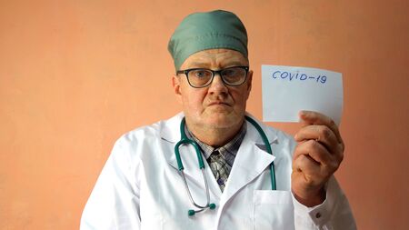 Medical doctor of Caucasian Ethnicity, in white coat, is holding sheet of paper with inscription Cowid-19 and warns of the danger of epidemic of coronavirus. Copy space for text. Close-up. Indoors.の写真素材