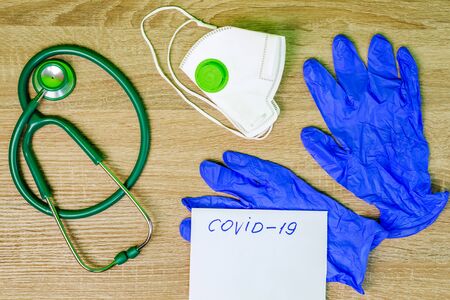 Respiratory mask, green stethoscope and rubber medical gloves, lie on wooden table. Inscription Covid-19 in frame.  Concept of medicine and healthcare. Copy space for text. Top view. Close-up. Indoors.の写真素材