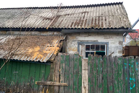 Facade of abandoned rickety brick house in village behind green wooden fence. Close-up. Outdoors.のeditorial素材