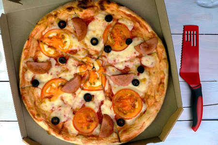 Fresh round pizza with tomatoes, sausage, mozzarella and olives in cardboard box. Pizza cutter and pizza spatula. Tasty unhealthy fast food. close-up.の写真素材