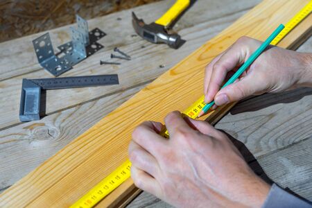 Male carpenter notes with pencil using measuring tape on the board, marks for saw cut, male hands with pencil and measuring tape close-up on wooden board.の写真素材
