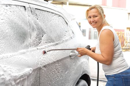 Smiling woman from a high-pressure hose applies a cleaner on the car at a car washの写真素材