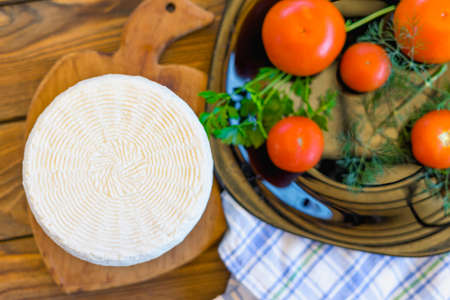 Cheese head on wooden board on wooden table served with tomatoes and fresh greens and towel. Homemade cheese. Food concept. Selective focus.の写真素材