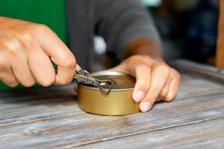 Mans hands opening a tin can on old wooden table with an old can opener with a metal handle. Canned...