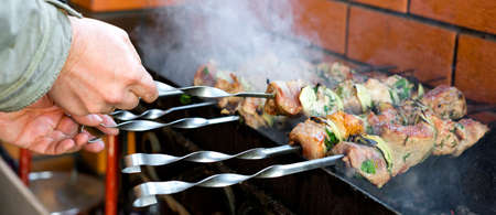 On the barbecue, a lot of meat is roasted on skewers, the chef turns over his shish kebabs. A mans hand turns, fries tasty meat of pork on skewers. Shallow depth of field.の写真素材