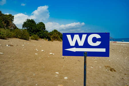 Direction to the toilet on the beach. WC - inscription on signboard toilet hanging on a pole, a sea, beach, green trees and sky background. Sunny summer day.の写真素材