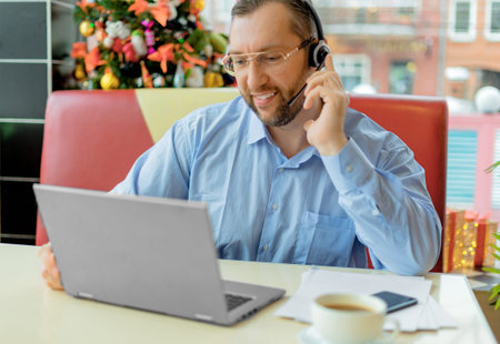 A middle-aged caucasian man with a headset in eyewear talking via video link with smile. He works...