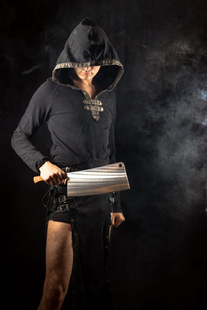 Young man posing in hooded gothic clothes with big cleaver. Royalty free stock photo.