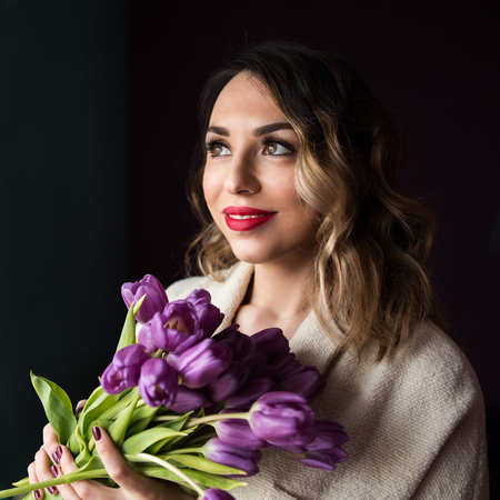 beautiful young woman looking out the window and holding flowers in the faceの写真素材