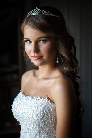 Beautiful young bride with wedding makeup and hairstyle by the window, newlywed woman final preparation for wedding. Happy Bride waiting groom.の写真素材