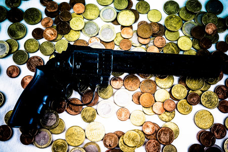 A gun is on top of a pile of coins. The coins are of different sizes and colors, and they are scattered around the gun. Concept of danger and violence, as the gun is a symbol of potential harm, real imageの写真素材