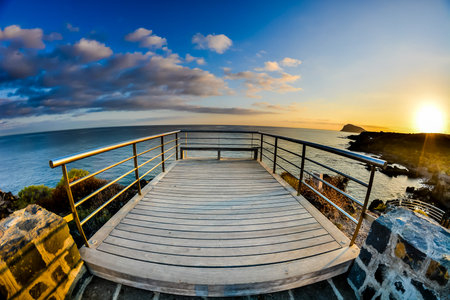 A wooden deck overlooking the ocean with a view of the sun setting. The sky is blue with some clouds, and the water is calm. The scene is peaceful and serene, with the sun setting in the distanceの写真素材