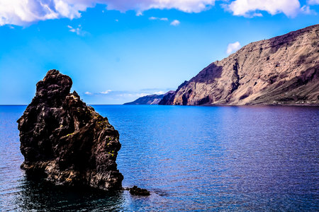 A large rock sits on the shore of a calm blue ocean. The rock is surrounded by water and the sky is clear and blue. The scene is peaceful and sereneの写真素材