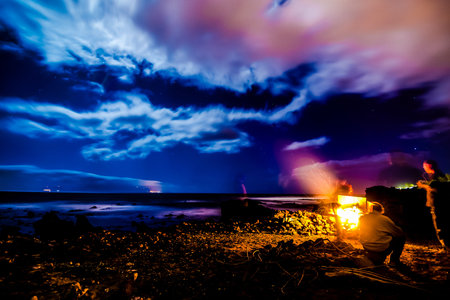 A man sits by a fire on a beach at night. The sky is cloudy and the water is calm
