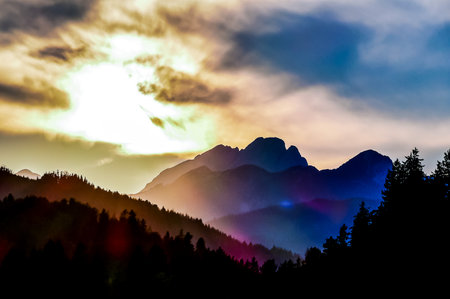 The mountains are covered in trees and the sky is a beautiful mix of colors. The sun is setting, casting a warm glow over the landscape. The scene is peaceful and serene, with the mountainsの写真素材