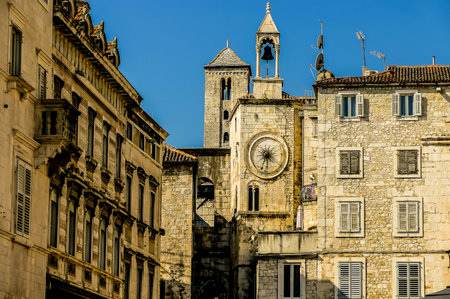 A city street with a large clock tower in the middle. The clock tower is surrounded by buildings and has a bell on top. The buildings are old and have a lot of windows. Scene is peaceful and nostalgicの写真素材