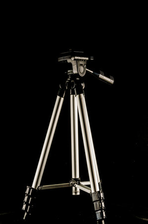 A tripod is standing on a black background. The tripod is silver and has three legs. The tripod is not in useの写真素材