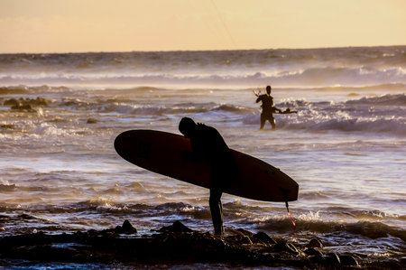 A surfer is walking away from the water with his surfboard. The sky is orange and the water is calmの写真素材