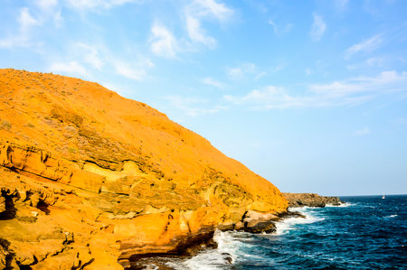 A rocky cliff overlooks the ocean. The sky is clear and blue, with a few clouds scattered throughout. The water is calm and the waves are small. The scene is peaceful and serene, with the oceanの写真素材