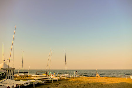 A beach with a few boats and a person standing on the sand. The sky is blue and the sun is settingの写真素材