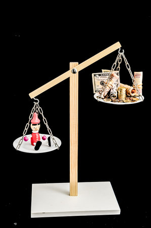 A wooden scale with a figurine on one side and money on the other. The figurine is wearing a red hat and is sitting on a platform. The money is in the form of coins and bills. The scale is balanced.の写真素材