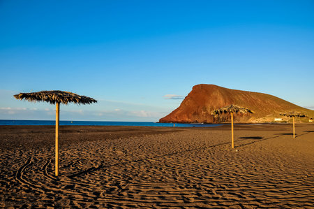 A beach with two umbrellas and a mountain in the background. The sky is blue and the beach is emptyの写真素材