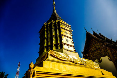 A tall yellow building with a statue of Buddha on top. The sky is blue and clear. The building is surrounded by treesの写真素材