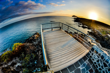 A beautiful view of the ocean with a wooden boardwalk leading to the water. The sun is setting, casting a warm glow over the sceneの写真素材