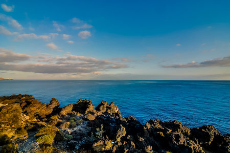 The ocean is calm and blue, with a few clouds in the sky. The rocks on the shoreline are large and jagged, creating a rugged and wild landscape. The scene is peaceful and serene, with the waterの写真素材