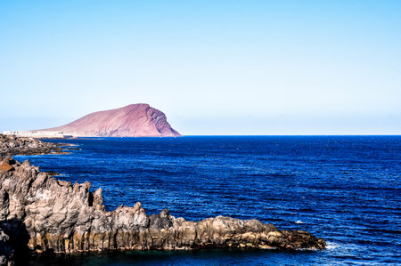 A rocky shoreline with a large mountain in the background. The ocean is calm and blue. The scene is peaceful and sereneの写真素材