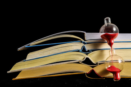 A book with a glass of sand on top of it. The sand is falling into the glass, creating a sense of time passing by. The scene is set on a black background, which adds to the overall mood of the imageの写真素材