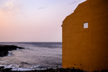 A building with a window overlooking the ocean. The sky is pink and the water is calm. The building is brown and the window is openの写真素材