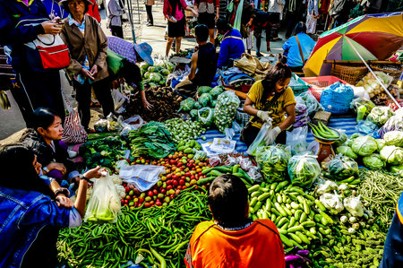 A group of people are gathered around a vegetable stand, with a variety of produce on display. The atmosphere is lively and bustling, with people browsing and shopping for fresh produceの写真素材