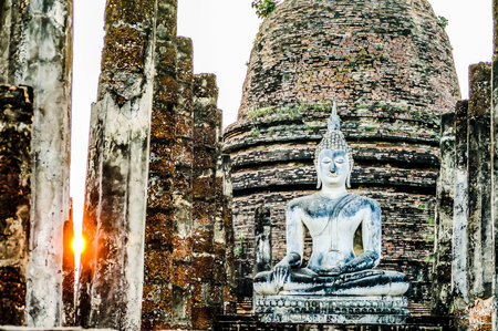 A statue of Buddha sits in front of a building with a large dome. The sun is setting behind the statue, casting a warm glow on the scene. Concept of peace and serenityの写真素材