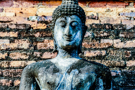 A statue of a Buddha with a stone face stands in front of a brick wall. The statue is old and has a stone face, giving it a sense of timelessness and wisdomの写真素材
