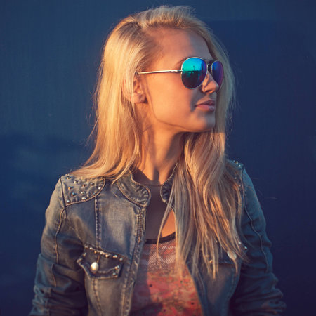 Outdoor summer closeup portrait of young stylish fashion glamorous smilling woman or girl in  sunny day on street jeans jacket and sunglasses standing near blue wallの写真素材