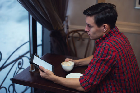 Handsome young man reading news using tablet while drinking coffee in a cafe sitting backside to the...