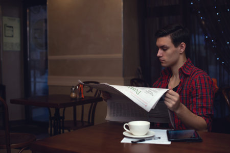 YOung hipster reading newspaper in a cafe during lunch with cup ot tea coffee