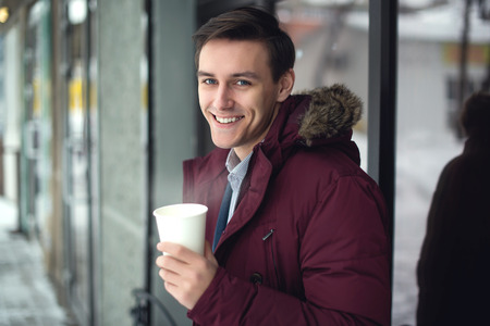 Young businessman in a suit and winter coat is drinking hot coffee at cold street with steam and bright smile on his faceの写真素材