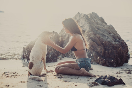 Portrait of beautiful happy young woman sitting playing with her pet dog sunset at the beach on the sea shore twilight sky as background.の写真素材